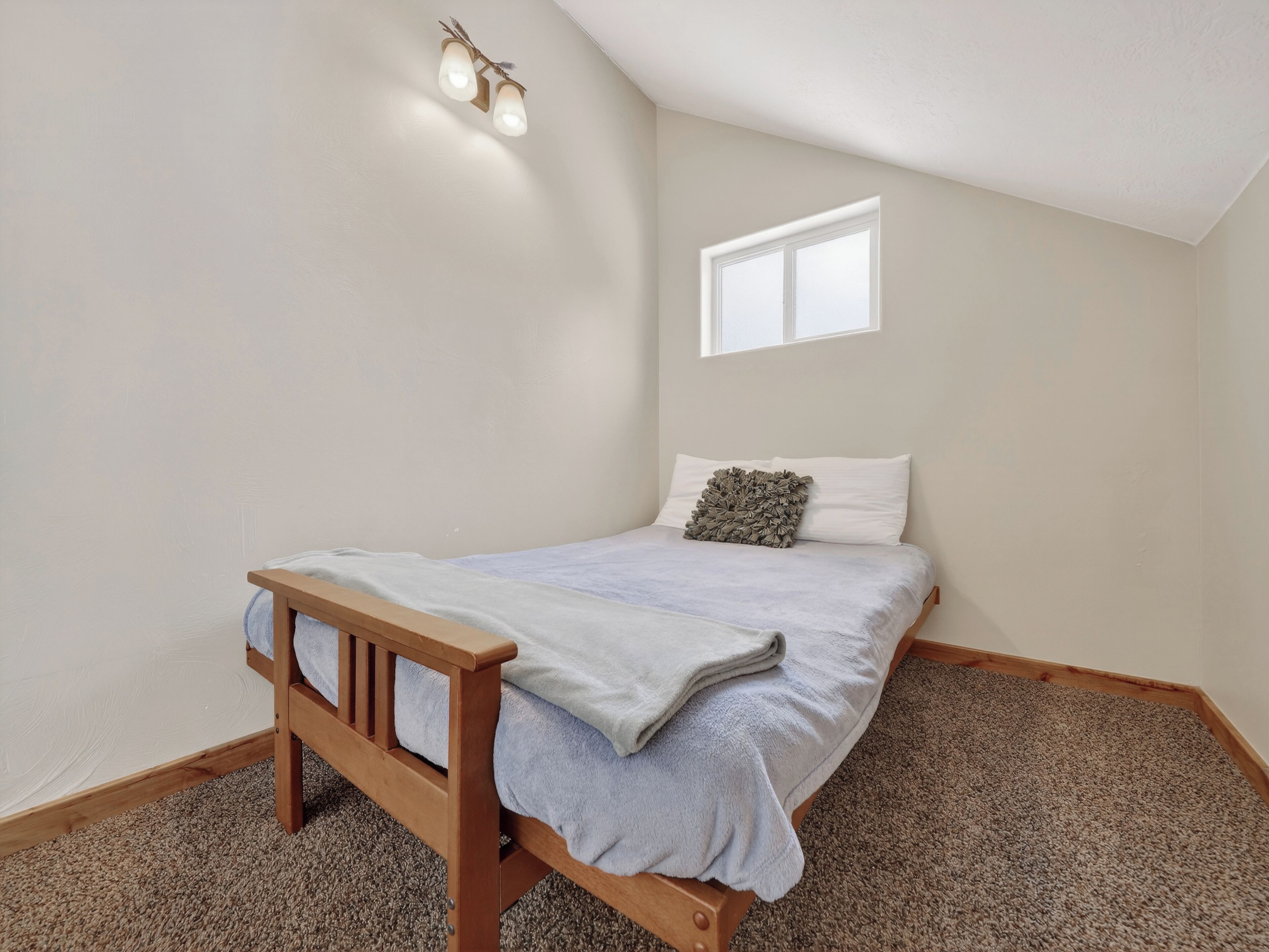 A bedroom with a single bed, a wooden bench, a small window, and a wall-mounted light fixture.