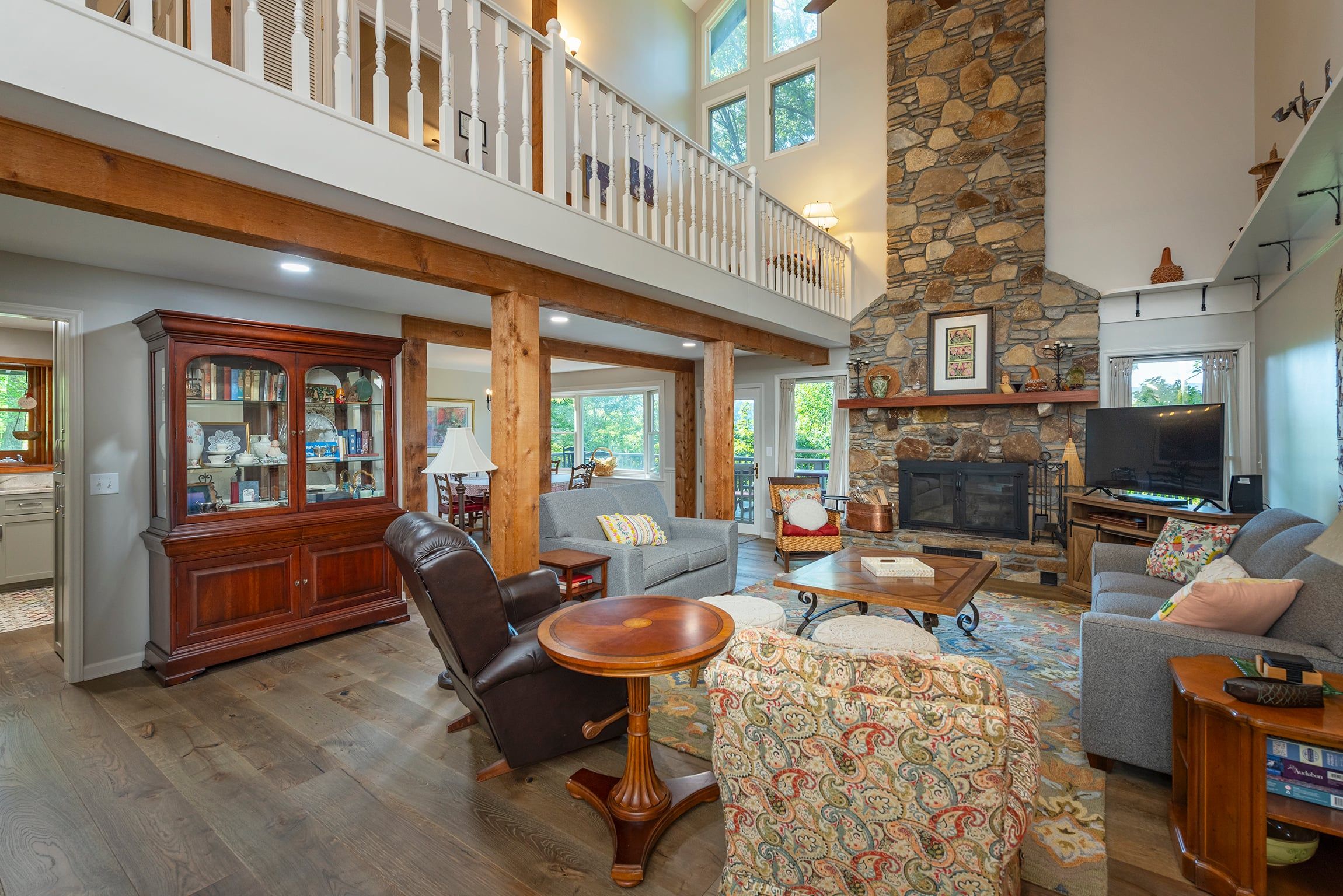 A spacious living room with a high ceiling, a stone fireplace, and a balcony with white railings.