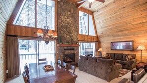 Un intérieur de chalet chaleureux avec une cheminée en pierre, des murs en bois, un coin repas et un salon doté d’un canapé et d’une télévision.