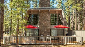 Une cabane avec une cheminée en pierre, deux grandes fenêtres surmontées de parasols rouges et une terrasse dotée d’une balustrade blanche.