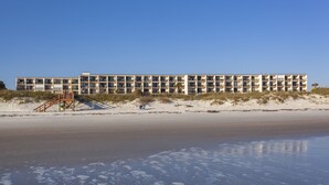 A multi-story hotel with a flat roof, located on a sandy beach with a staircase leading to the entrance.