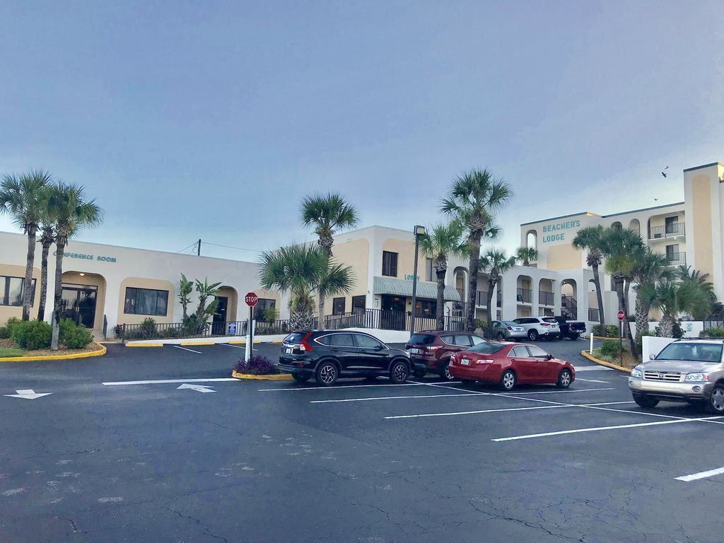 Un estacionamiento con varios coches, palmeras y edificios que señalan un salón de conferencias y una cabaña frente al mar.