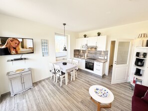 Cozinha compacta com armários brancos, micro-ondas e fogão. Há uma mesa de jantar com cadeiras e uma pequena mesa com um prato. Uma TV está instalada na parede, e há uma janela com vista para edifícios.