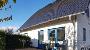 A white building with blue doors and windows, a tiled roof, and a paved patio area with outdoor furniture. - Hus at Zickenbarg - Apartment Hus at Zickenbarg (Zempin)