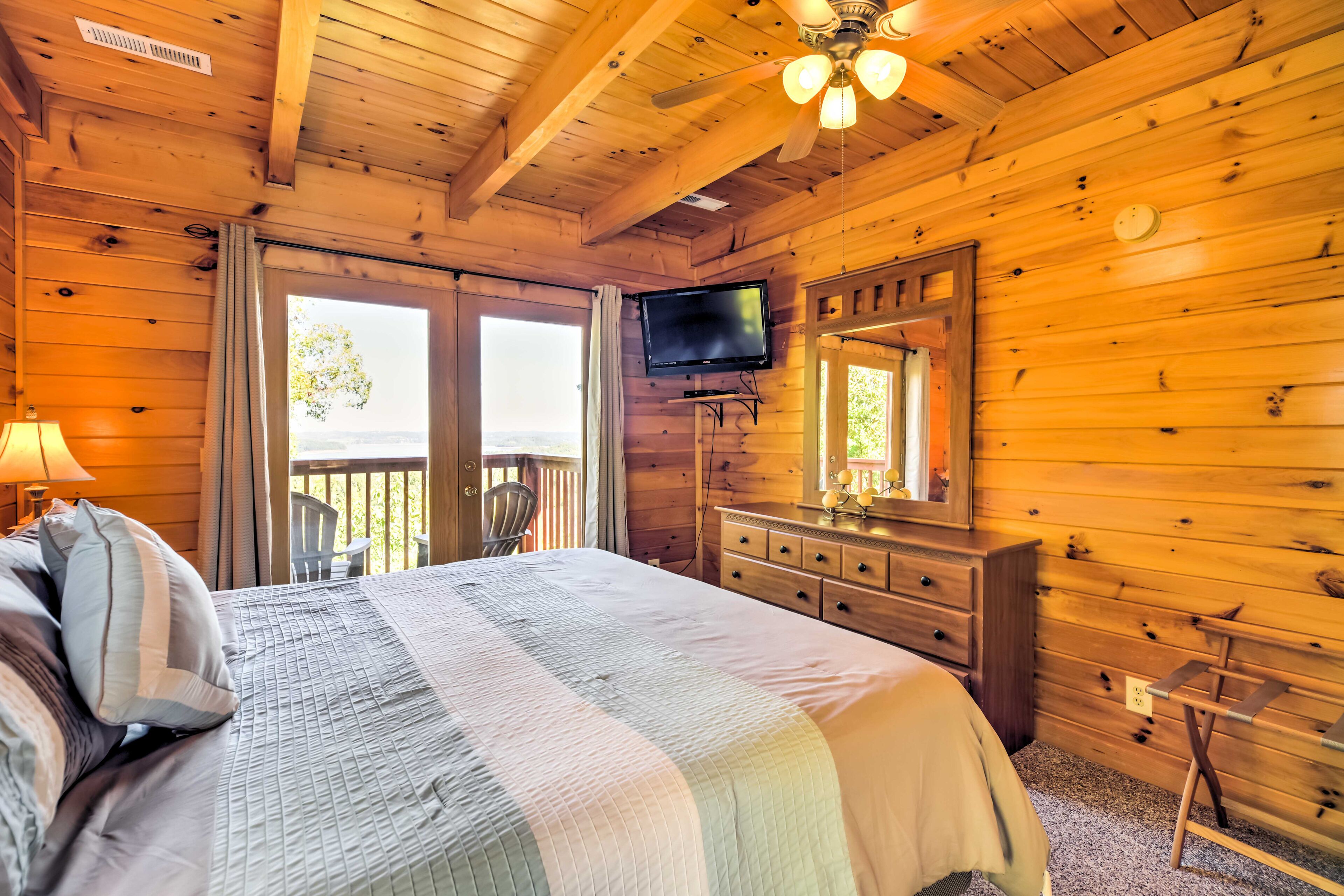 Un dormitorio de cabaña de madera con cama, cómoda, televisor y balcón con una silla.