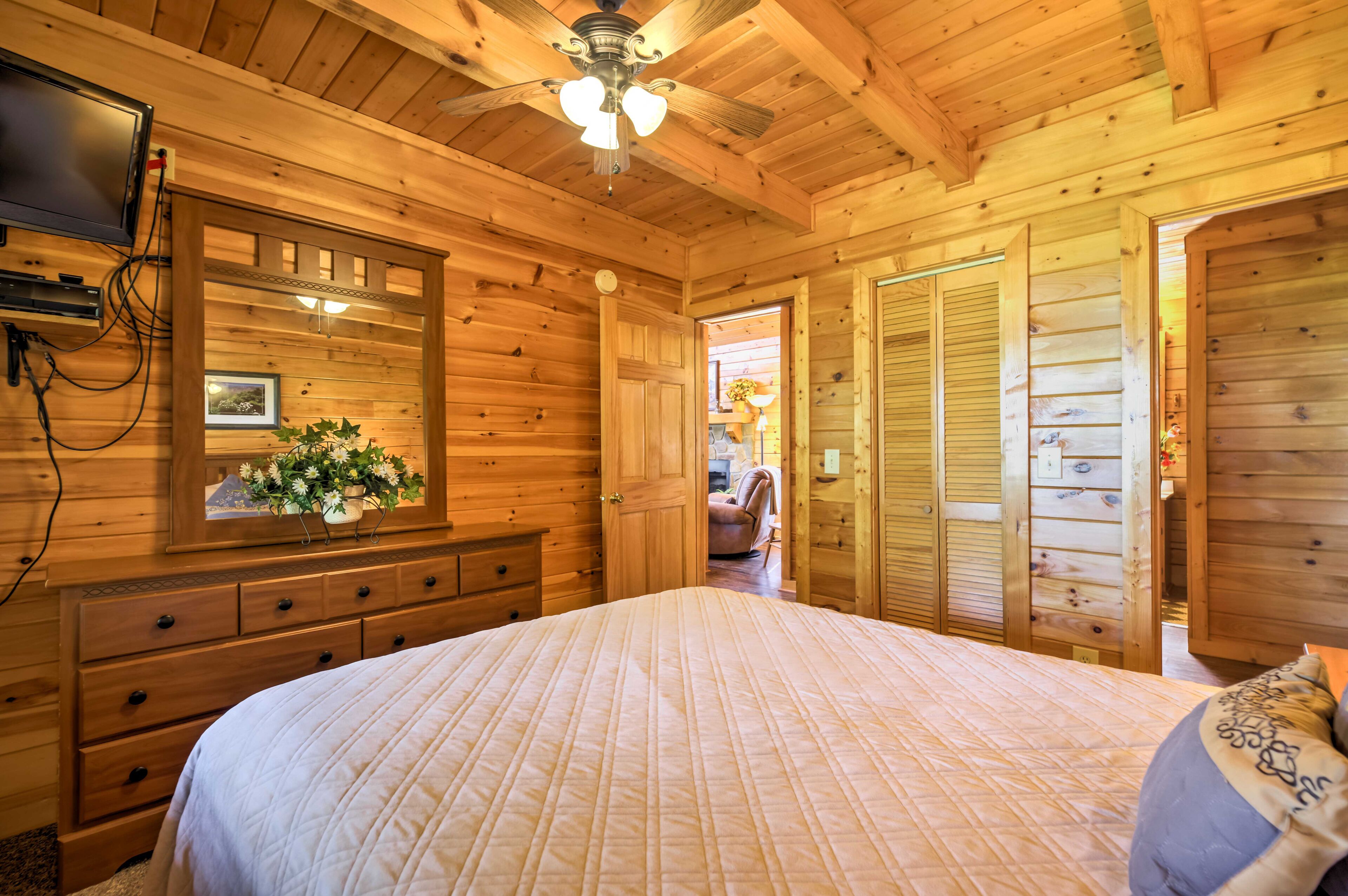 A wooden cabin bedroom with a bed, dresser, TV, and a ceiling fan.