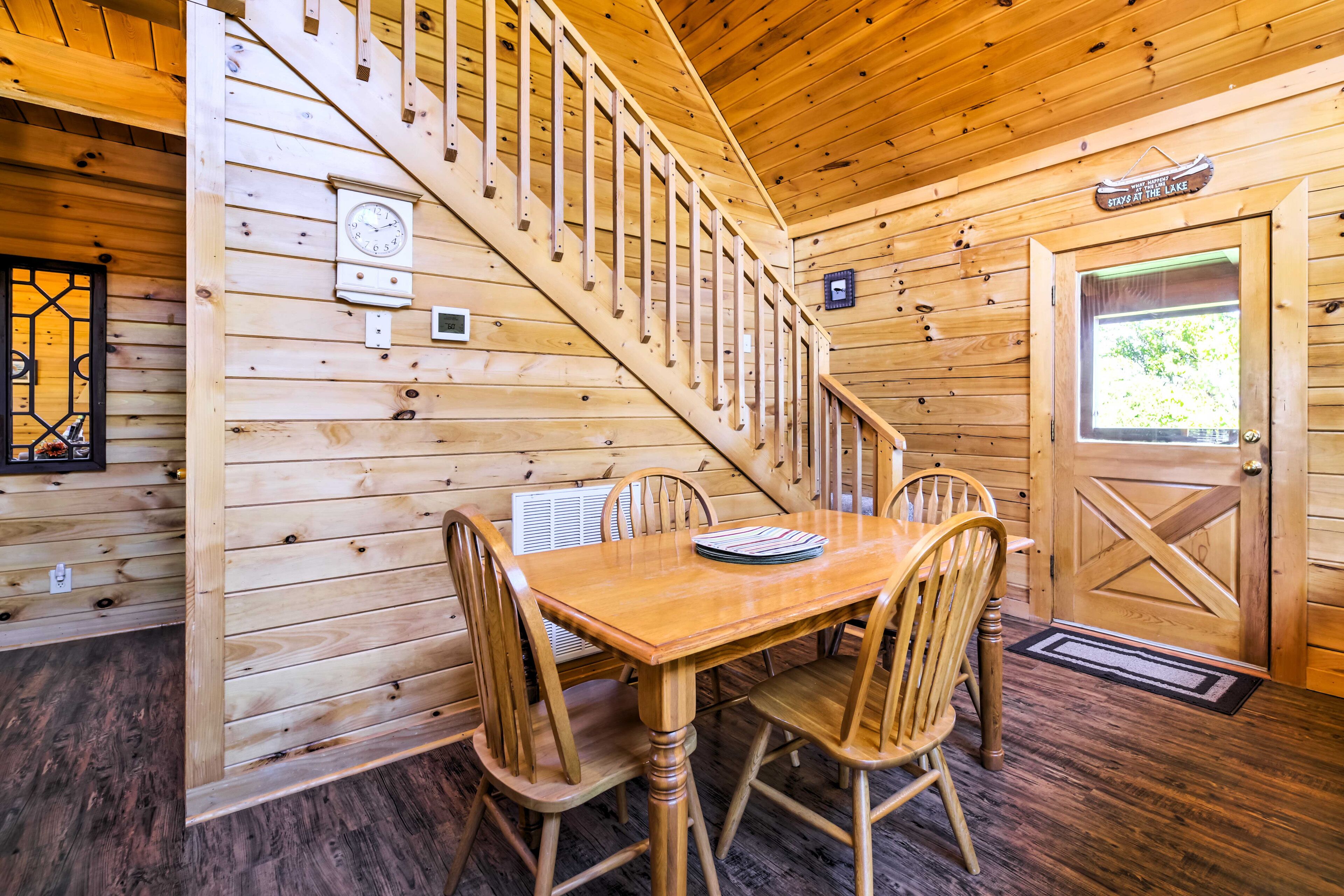 A wooden cabin interior with a dining table, four chairs, a staircase, a clock, and a door with a window.