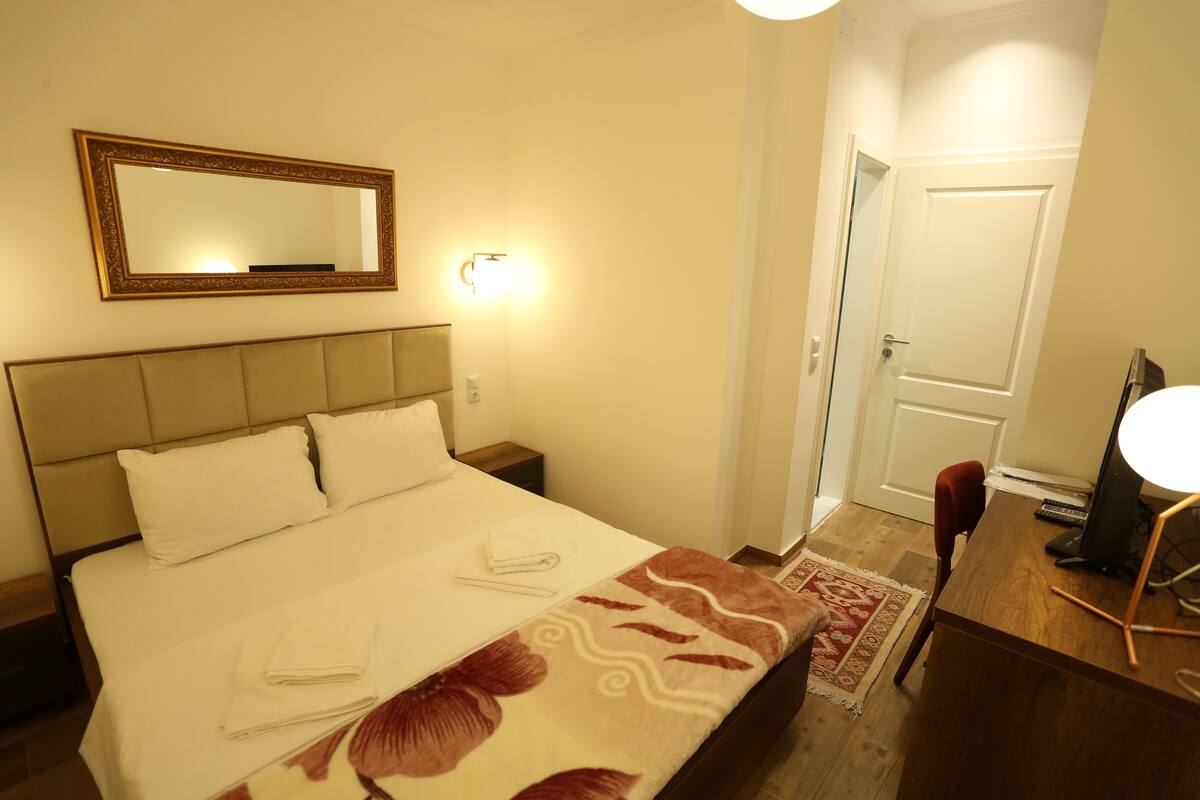 A hotel room with a bed, a desk, a chair, a desk lamp, a computer, a mirror, and a door.