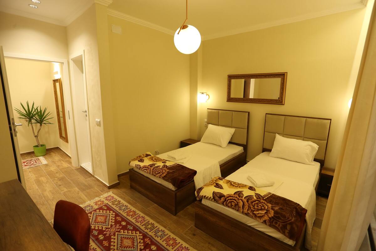 A hotel room with two beds, a small table, a chair, a potted plant, and a mirror on the wall.