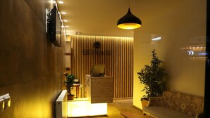 A reception area with a wooden reception desk, a sofa, a potted plant, and a hanging light fixture. - Hotel Edi Imperial (Prizren)