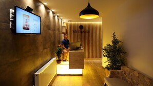 A reception area with a wooden reception desk, a television mounted on the wall, and a potted plant. - Hotel Edi Imperial (Prizren)