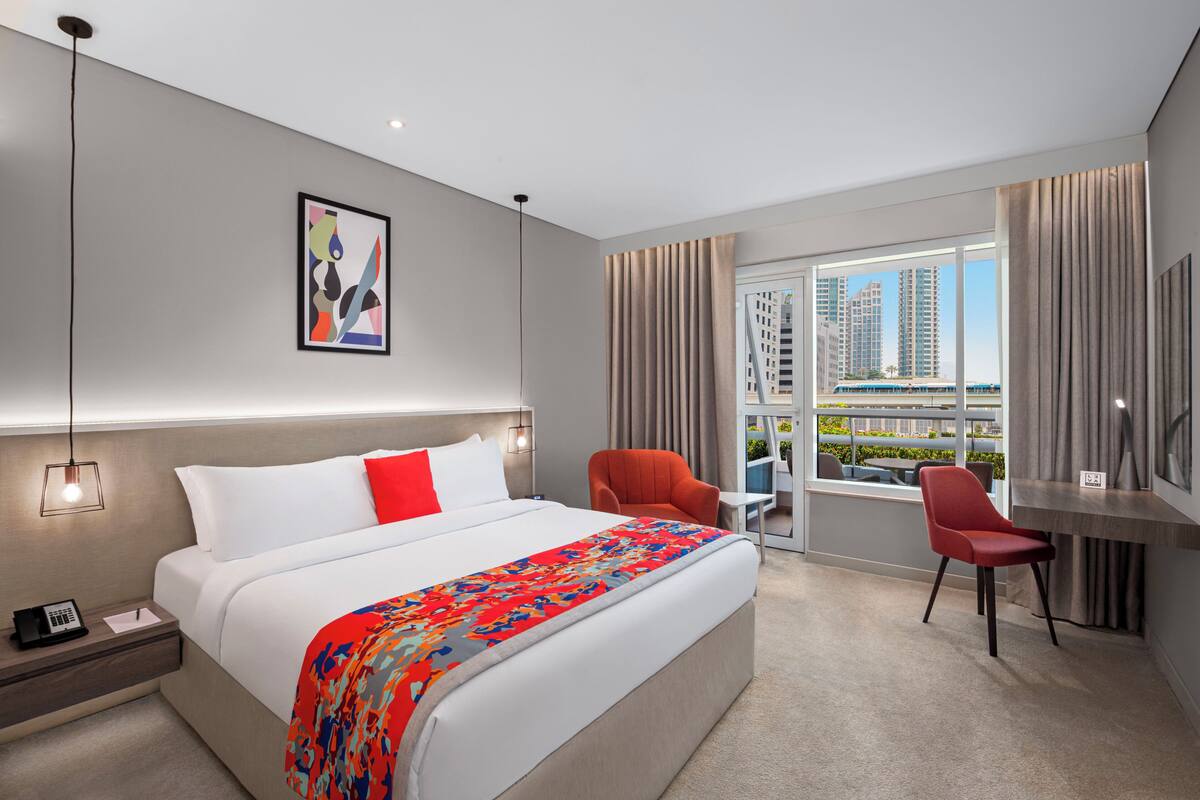 A modern hotel room with a large bed, a desk, and a chair. There is a window with a view of buildings.