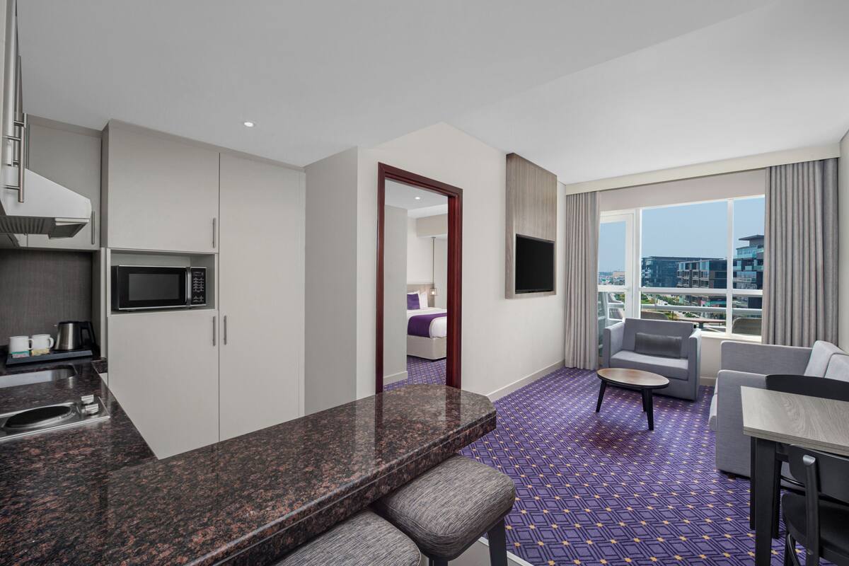 A modern hotel room with a kitchen area, a seating area with a sofa and armchairs, and a large window offering a city view.