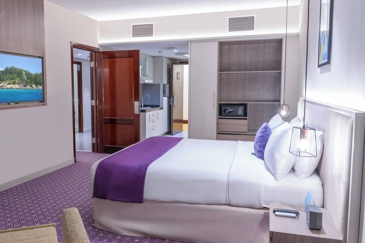 A hotel room with a bed, a desk, a chair, a TV, and a view of a beach.