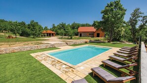 A rectangular swimming pool with a stone border, surrounded by a lawn and a stone wall. There are wooden lounge chairs with cushions along the pool's edge. - Pet friendly home in Perusic (Perusic)