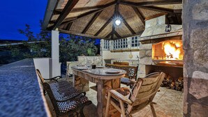 An outdoor patio with a stone table and chairs, a stone fireplace, and a thatched roof structure.