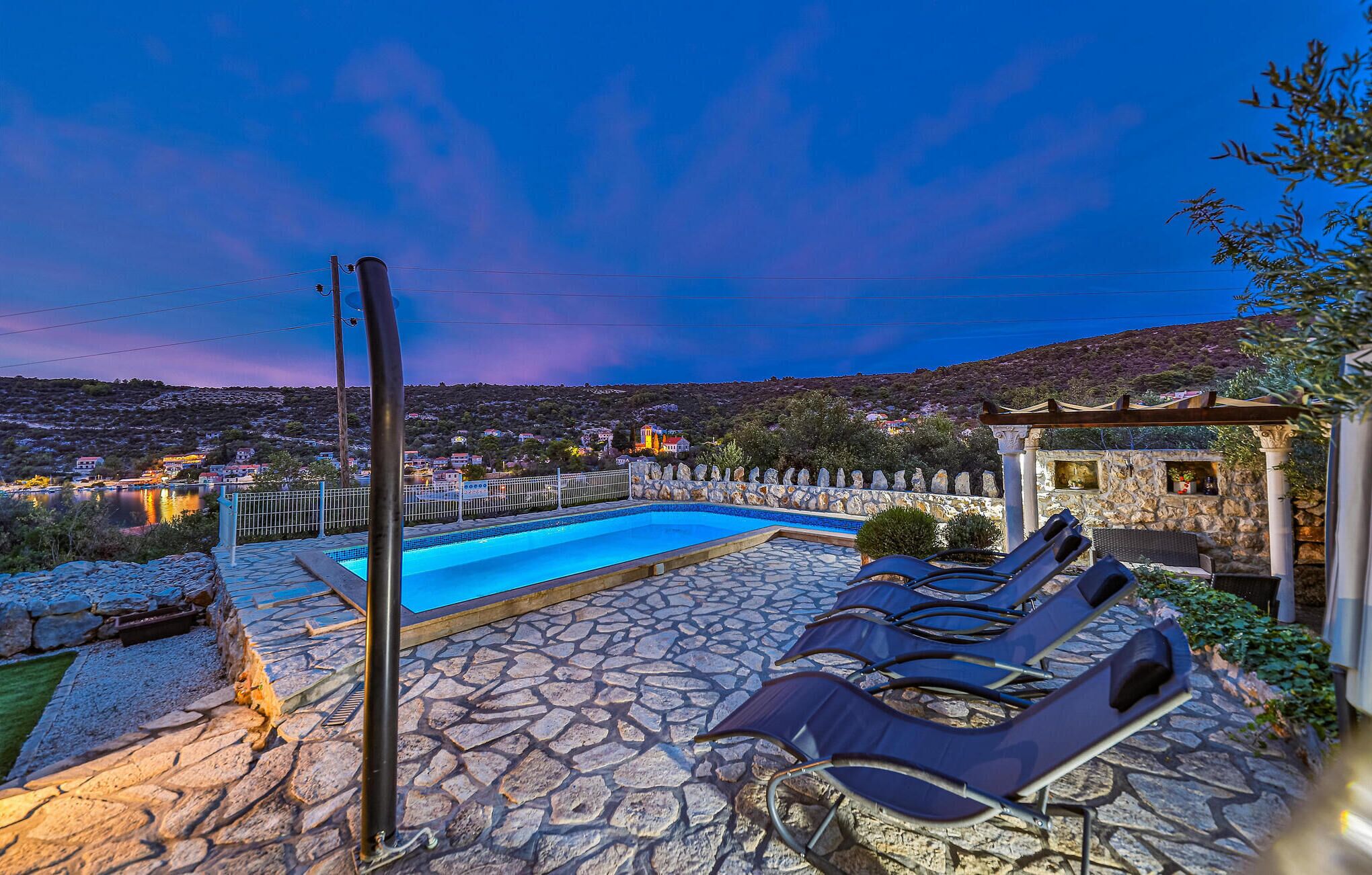 Una piscina con sillas de descanso, un patio de piedra y vistas a un pueblo en la colina al anochecer.