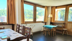 A room with a dining table, chairs, a small table with a blue tablecloth, and a lamp. There are large windows with curtains, a radiator, and a view of greenery outside. - Large apartment for 8 in Plagne Centre, balcony, pets allowed (La Plagne)