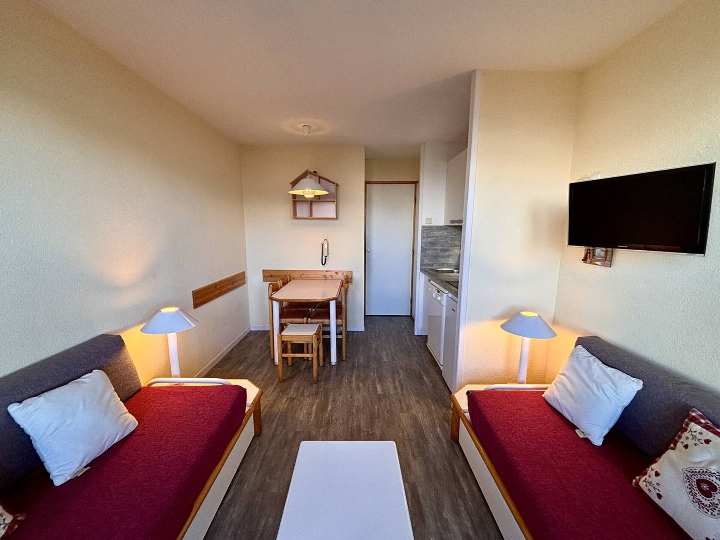 A compact hotel room with two single beds, a small dining table, a kitchenette, and a TV mounted on the wall.