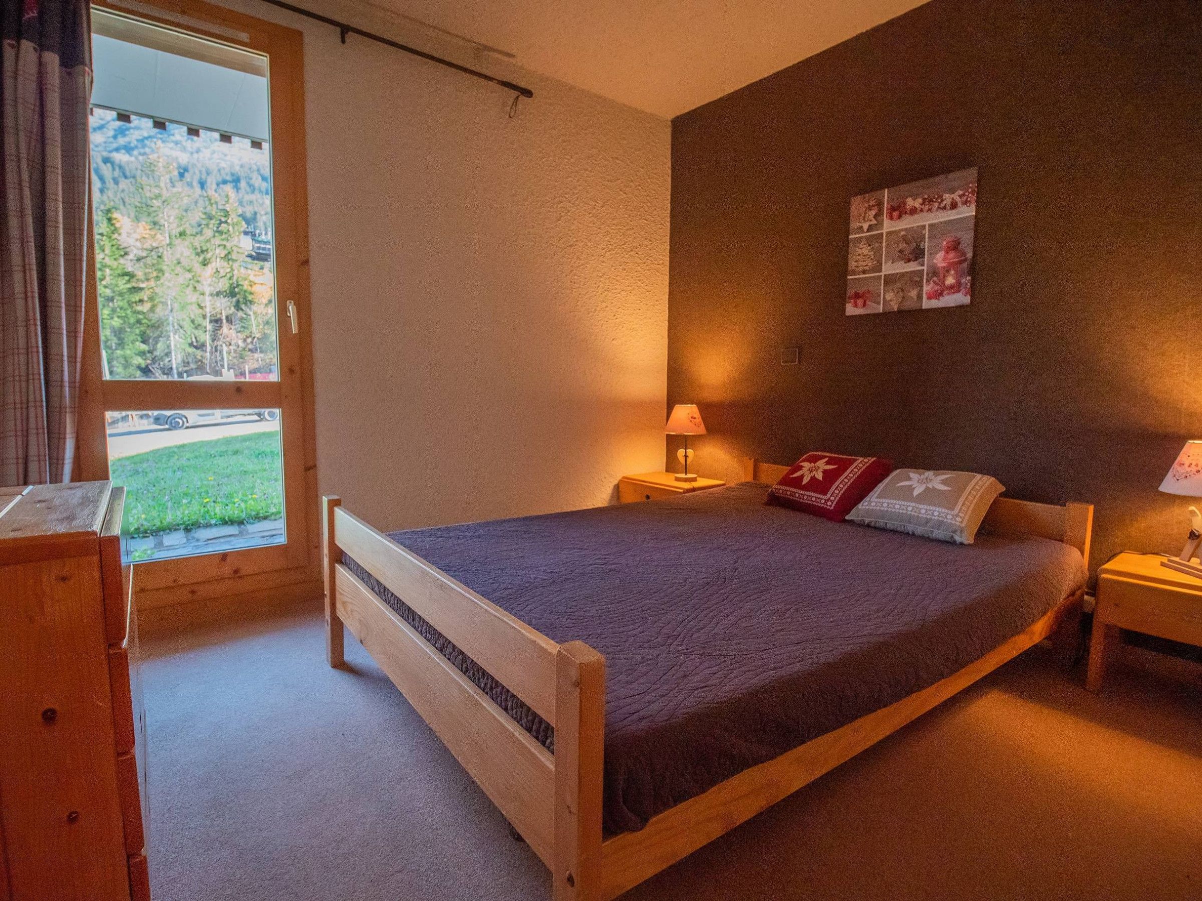 Ein Schlafzimmer mit einem hölzernen Bett, einem Nachttisch mit Lampe, einem Fenster mit Blick auf Bäume und einer Wand mit einem gerahmten Bild.