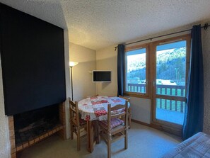 A cozy room with a fireplace, a table with chairs, a television, and a large window with a view of a deck and trees. - 1-bedroom in central Valmorel, close to shops and lifts (Valmorel)
