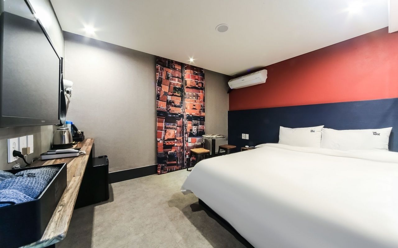 Hotellihuone, jossa on suuri sänky, työpöytä tuolilla, pieni pöytä, ilmastointilaite ja seinä, jossa on värikäs, abstrakti kuvio.