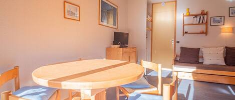 Ein gemütliches Zimmer mit einem Holzesstisch und Stühlen, einem Fernseher auf einer Holzkommode und einem Sofa mit Kissen.