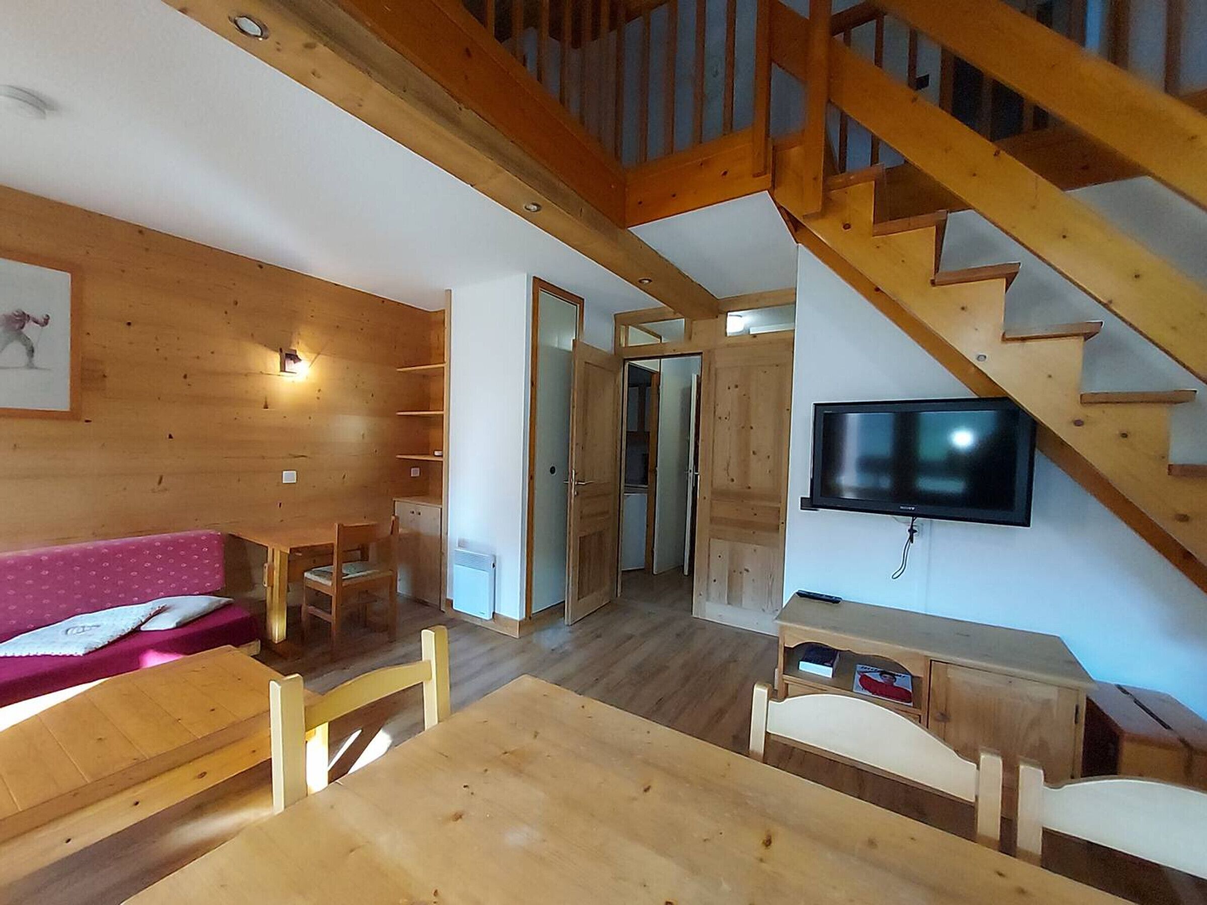 A cozy wooden interior with a staircase, a TV mounted on the wall, and a dining area with a table and chairs.