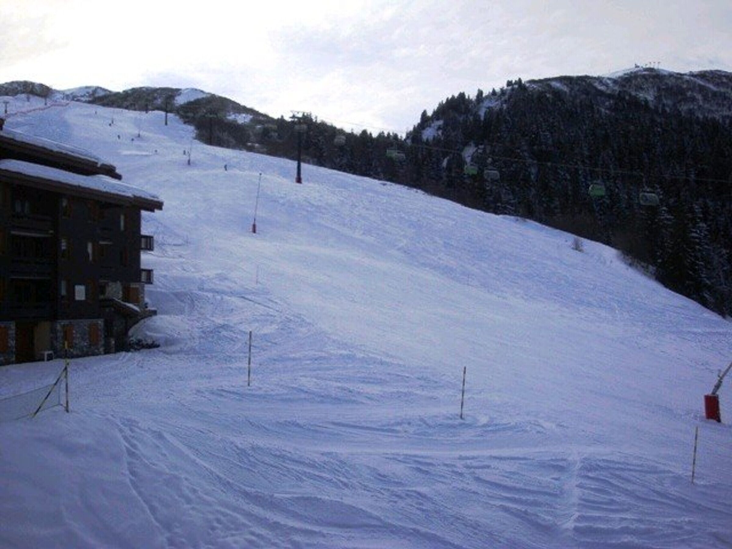 A snow-covered ski slope with a building on the left and a forested hill in the background.