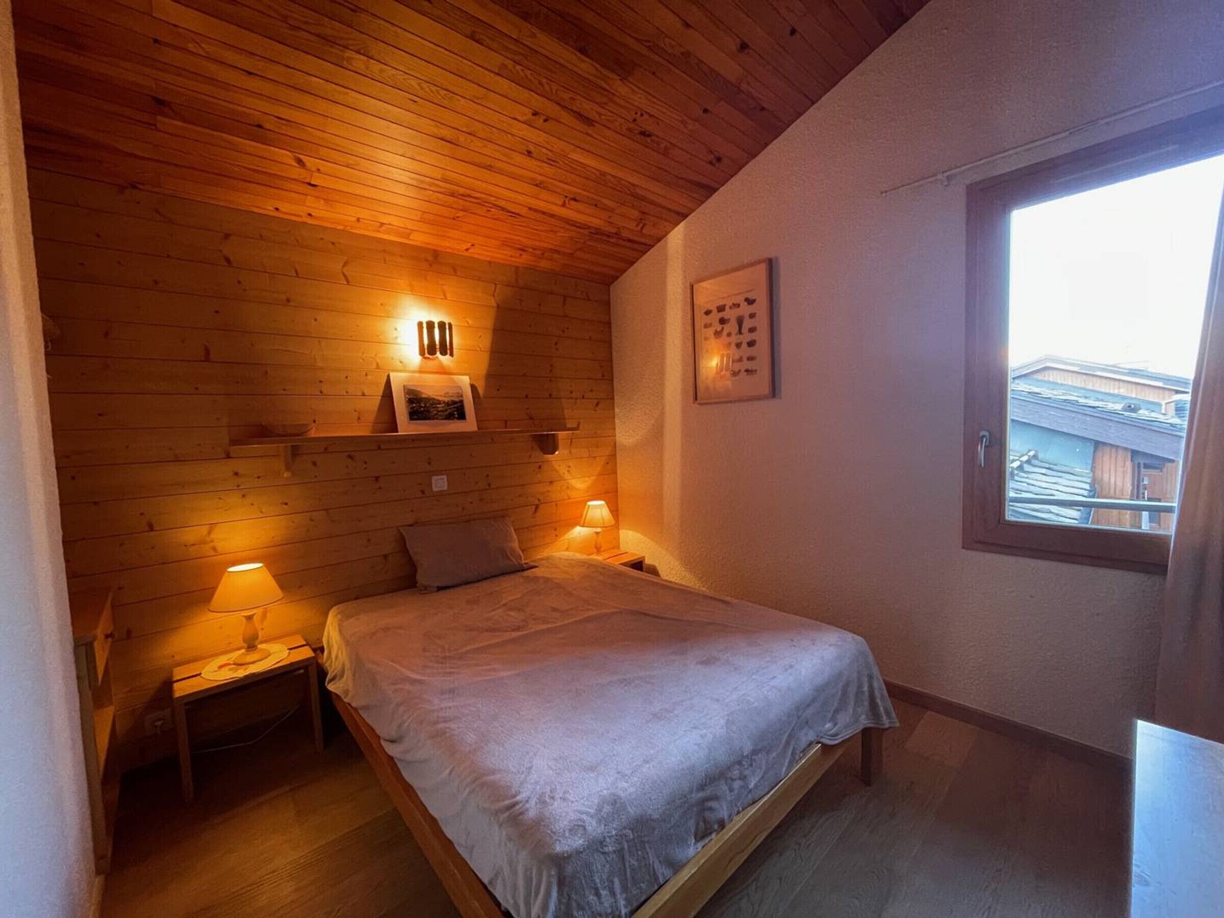 A cozy bedroom with a wooden ceiling, a single bed, two bedside lamps, and a window with a view of buildings outside.