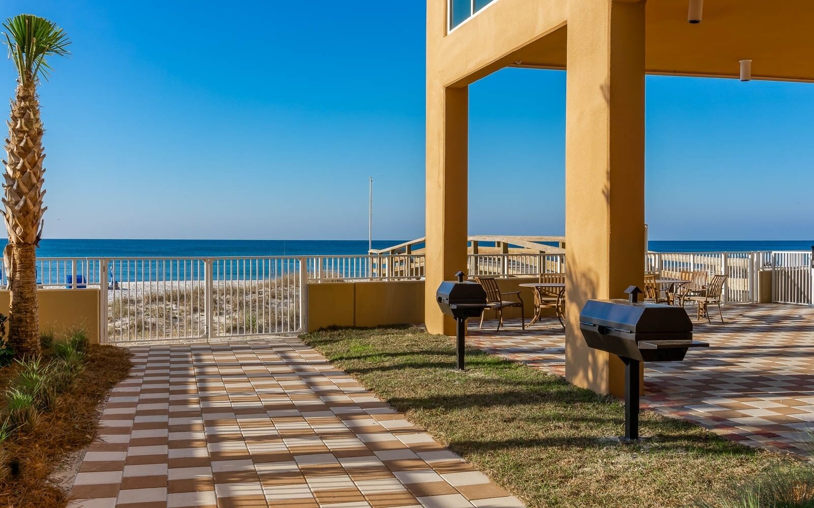 Un espace patio avec un chemin pavé menant à un coin salon aménagé avec du mobilier d’extérieur, un barbecue et une vue sur la plage et l’océan.