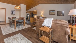 A spacious living area with a wooden ceiling, a brown leather sofa, a dining table with chairs, and a kitchen in the background. - Condo w/beautiful views, fireplace & community pool/hot tub - Cozy Hidden Village Retreat (Big Sky)