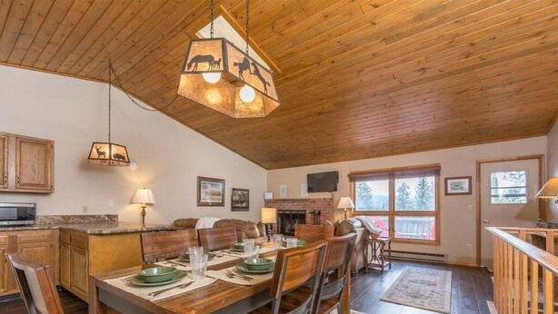 A spacious dining area with a wooden table and chairs, a hanging light fixture, and a fireplace in the background. - Condo w/beautiful views, fireplace & community pool/hot tub - Cozy Hidden Village Retreat (Big Sky)
