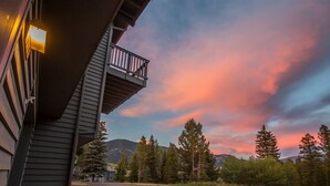 A building with a balcony overlooking a parking area and a mountainous landscape during sunset. - Condo w/beautiful views, fireplace & community pool/hot tub - Cozy Hidden Village Retreat (Big Sky)