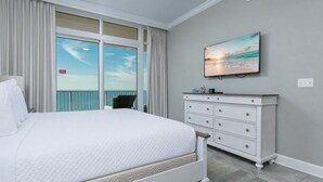 Habitación de hotel con cama, cómoda, televisor montado en la pared y vistas al océano a través de amplias ventanas.