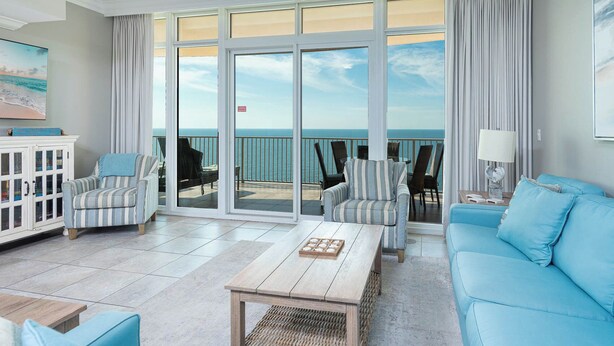Amplia sala de estar con un gran ventanal corredera que da a un balcón con vistas al mar. Hay dos sillones a rayas, un sofá azul y una mesa de centro de madera.