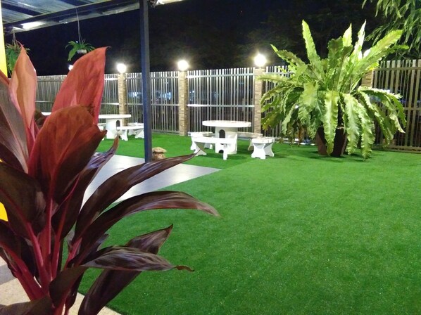 An outdoor patio area with artificial grass, white tables and chairs, and large potted plants. - Baan Wanchart Bangkok Residences (Bangkok)