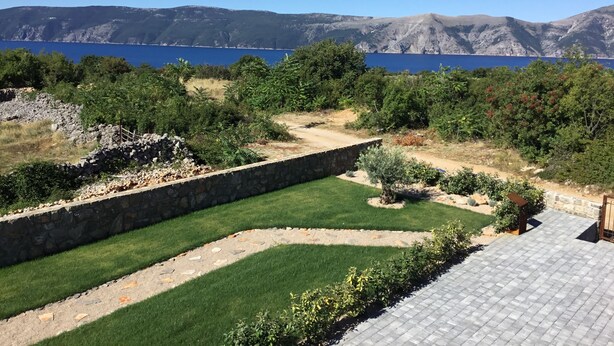 Een aangelegde tuin met een stenen muur, een geplaveid terras en uitzicht op een meer en bergen.