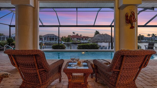 A poolside area with wicker chairs and a table, overlooking a body of water and a sunset view. - Gulf Access, Southern Heated Pool, Kayaks, Bikes, Tiki Hut - Villa Salty Shoreline - Roelens (Cape Coral)