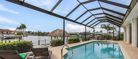 Piscina ao ar livre coberta com espreguiçadeiras, vista para a água e uma estrutura com telhado de palha ao fundo.