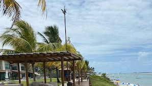 A wooden deck with outdoor seating, a covered area, and a view of the beach with umbrellas. - House in condominium by the sea, chicken harbor (high wall)<br> (Porto de Galinhas)