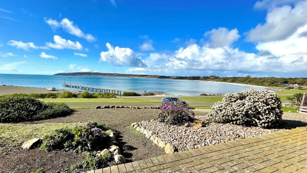 Un paysage côtier avec une jetée qui s’avance dans la mer, une zone herbeuse agrémentée d’un parterre de fleurs et un ciel dégagé.