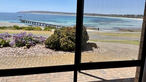 Une vue prise de l’intérieur d’un bâtiment, donnant sur une plage avec une jetée, un buisson et des fleurs violettes.