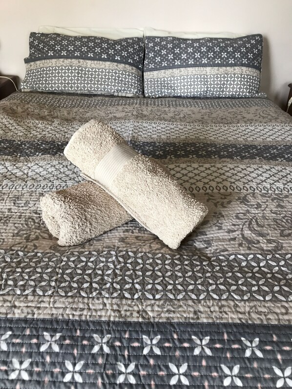 A bed with a patterned bedspread, two pillows, and a rolled-up towel.