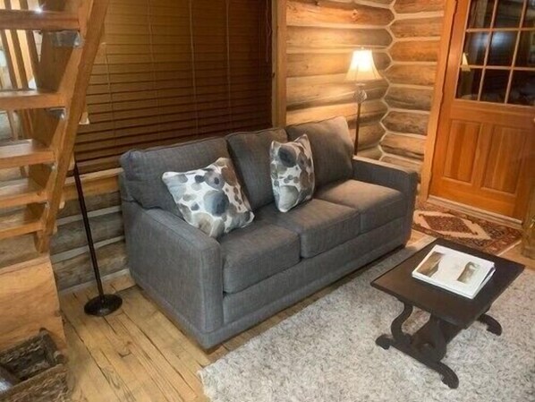 A grey sofa with two patterned cushions, a wooden staircase, a lamp, and a small table with a book. - A Log Cabin in the Woods (Mazama)