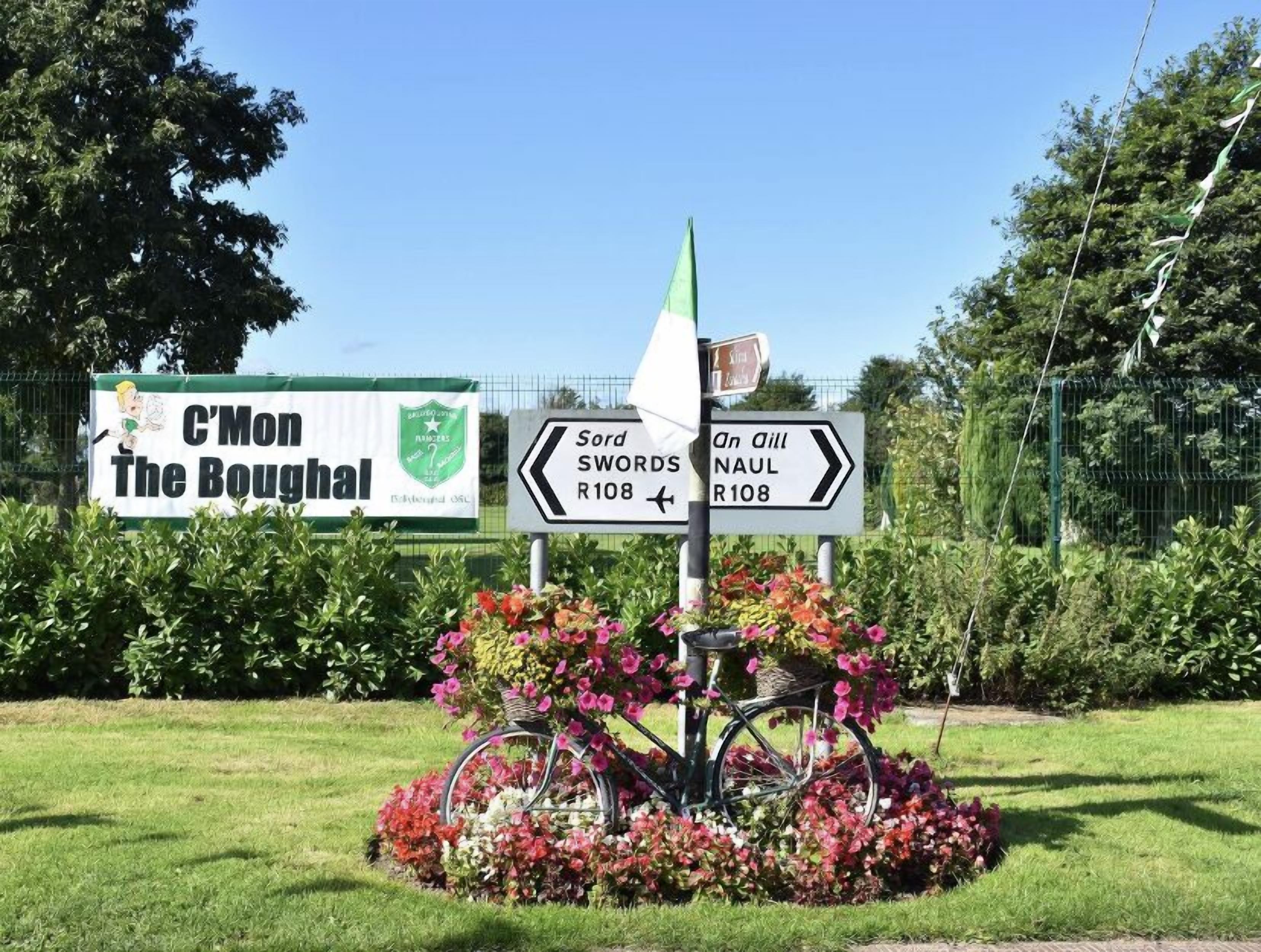 Een richtingwijzer met 'C'Mon The Boughal' en 'Sord Swords R108' naar links wijzend, en 'An Oll NAUL R108' naar rechts, omringd door bloemen en een fiets.