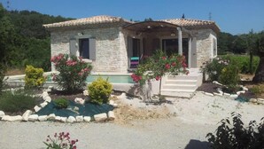 A stone house with a tiled roof, a porch, and a small garden with flowering plants.