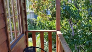 Une terrasse en bois avec une table en verre et deux chaises, donnant sur un jardin avec des arbres et une maison en arrière-plan.