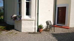 A building with a white exterior, a window with a flower box, a door with a red frame, and a chair outside. - The Edwardian Wing, 18th Century Oak with Wing inserts (Taynuilt)