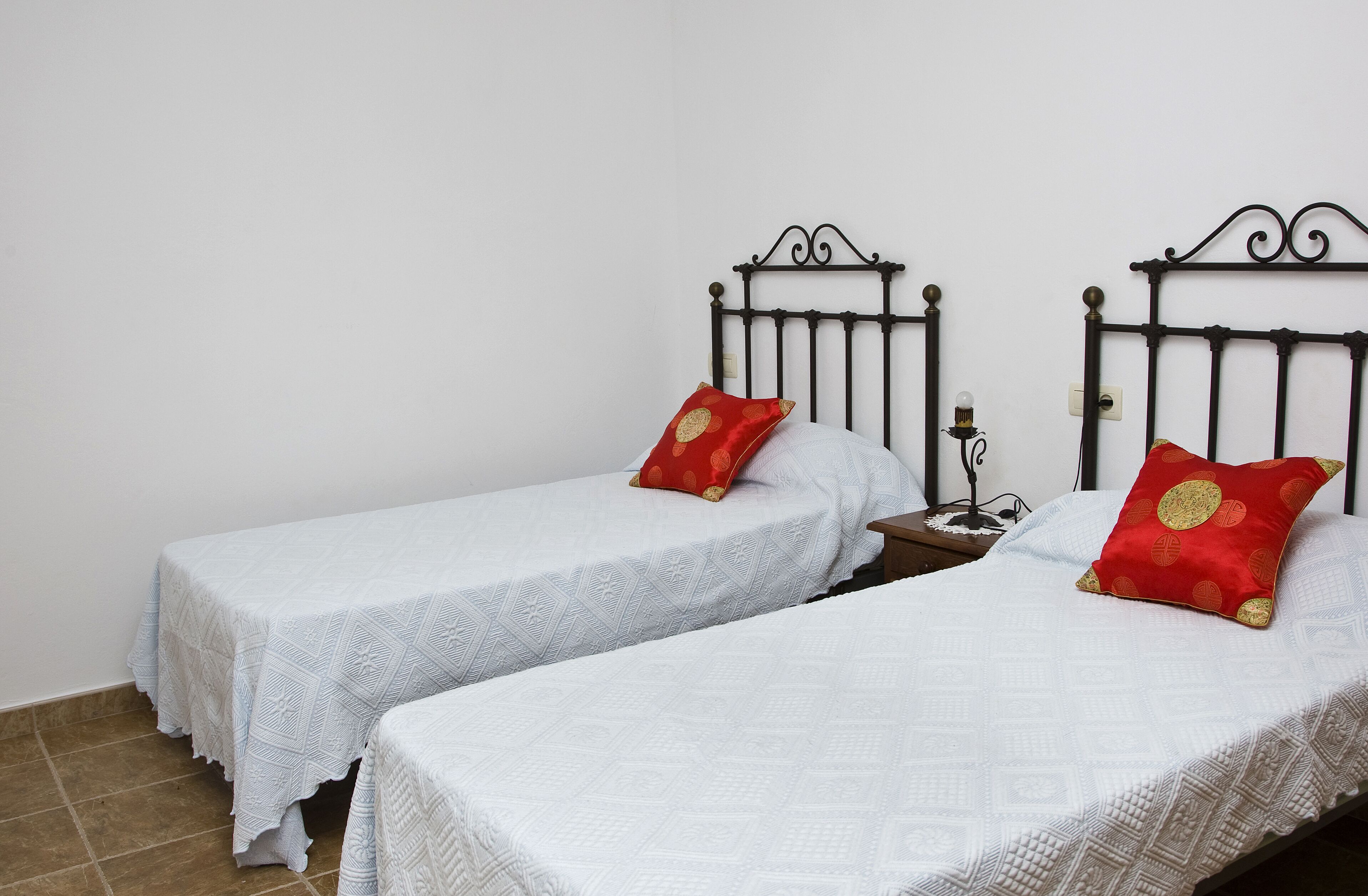 Deux lits simples avec du linge de lit blanc et des oreillers rouges, une table de chevet avec une lampe, et un mur blanc uni.